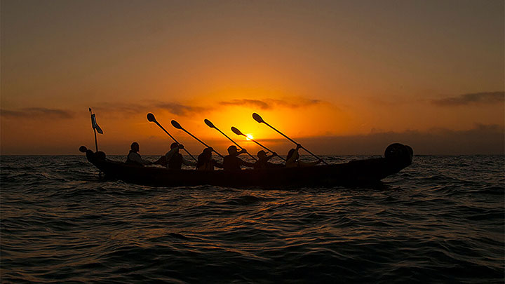 people holding paddles up in a boat on the ocean at sunset